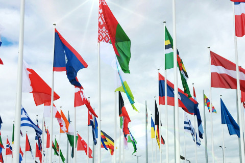 Auf dem Bild sind viele verschiedene Länderflaggen, die im Wind wehen. Im Hintergrund ist ein bewölkter Himmel.